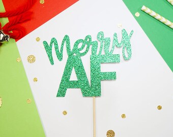 Könnte beinhalten: Ein grüner Glitzer-Kuchenaufsatz mit den Worten "Merry AF" auf einem Holzstab. Der Hintergrund umfasst rotes und grünes Papier, goldenes Konfetti und dekorative Strohhalme. Eine rote Schnur mit einer Glocke ist ebenfalls sichtbar.