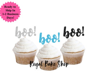 Könnte beinhalten: Drei Cupcakes mit weißem Zuckerguss, jeweils mit einem glitzernden "boo!" Tortenaufsatz in Silber, Blau und Schwarz. Die Cupcakes befinden sich in braunen Papierförmchen. Der Text "Royal Bake Shop" steht unten.