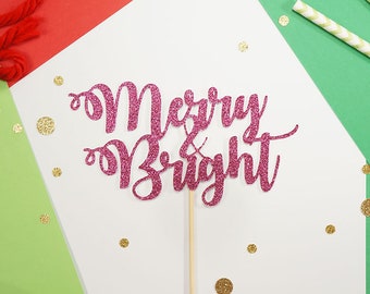 Könnte beinhalten: Ein pinkfarbener Glitzer-Kuchenaufsatz mit der Aufschrift "Merry & Bright" auf einem Holzstab. Der Aufsatz befindet sich auf einer weißen Oberfläche mit goldenem Konfetti, rotem Garn, einer silbernen Glocke und grünem und rotem Papier.