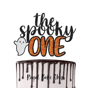 May include: A Halloween cake topper with the words "the spooky ONE" in black glitter script and orange glitter block letters. A white glitter ghost is on the left. The cake has chocolate frosting dripping down the sides.