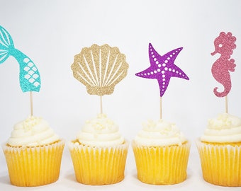 Könnte beinhalten: Vier Cupcakes, die mit Glitzer-Toppern verziert sind. Die Topper sind ein türkisfarbener Meerjungfrauen-Schwanz, eine goldene Muschel, ein lilafarbener Seestern und ein rosafarbenes Seepferdchen.