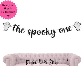 Könnte beinhalten: Ein Halloween-Banner mit dem Schriftzug "the spooky one" in schwarzem Glitzer, verziert mit zwei silbernen Geistern. Der Banner ist vor einem weißen Hintergrund platziert. Der Schriftzug "Royal Bake Shop" ist unten zu sehen.
