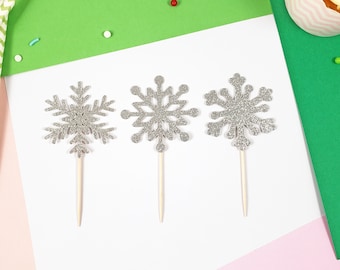 Könnte beinhalten: Drei silberne Glitzer-Schneeflocken-Kuchentopper auf weißen Stielen. Die Schneeflocken sind unterschiedlich groß und haben einen glitzernden Überzug.