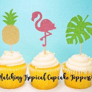 Puede incluir: Tres cupcakes con adornos de cupcake tropicales a juego. Los adornos son un flamenco rosa con purpurina, una hoja de monstera verde con purpurina y una pi&ntilde;a dorada con purpurina.