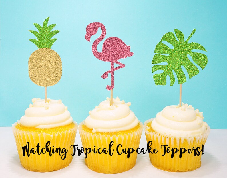 Puede incluir: Tres cupcakes con adornos de cupcake tropicales a juego. Los adornos son un flamenco rosa con purpurina, una hoja de monstera verde con purpurina y una pi&ntilde;a dorada con purpurina.
