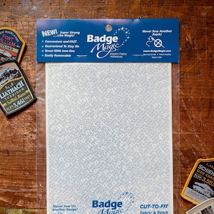 Può includere: Una confezione di adesivo istantaneo per tessuti Badge Magic, con la scritta "Cut-to-Fit Fabric & Patch Adhesive". La confezione è trasparente, con un'etichetta blu e bianca. Sono visibili toppe intorno alla confezione.
