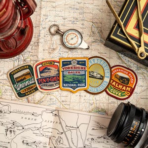 May include: A selection of Yorkshire Dales patches, a compass, and a vintage map. The patches display text and illustrations of local landmarks. A red lantern and a camera lens are also visible, suggesting a travel theme.