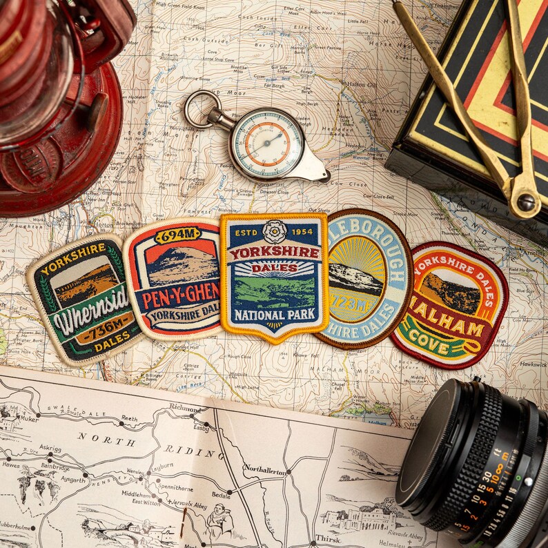 May include: A selection of Yorkshire Dales patches, a compass, and a vintage map. The patches display text and illustrations of local landmarks. A red lantern and a camera lens are also visible, suggesting a travel theme.