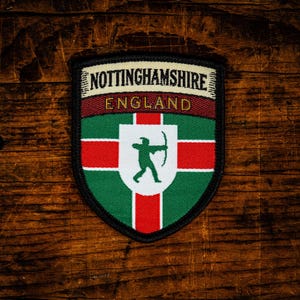 May include: A shield-shaped embroidered patch featuring the text NOTTINGHAMSHIRE and ENGLAND. The design includes a green and red cross background with a white central shield displaying a green silhouette of an archer aiming a bow and arrow.