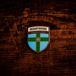 May include: A shield-shaped emblem with the words "DERBYSHIRE ENGLAND" at the top. The shield features a blue background with a green cross and a yellow floral design in the center. The emblem has a silver border.