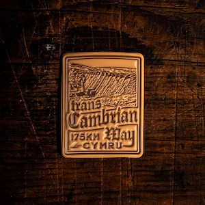 Pode incluir: Um alfinete retangular de metal cor de bronze com as palavras "trans Cambrian Way" e "175KM May CYMRU" em uma fonte decorativa. O alfinete apresenta uma cena de paisagem. O alfinete está sobre uma superfície de madeira escura.