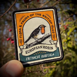 Könnte beinhalten: Gestickter Aufnäher mit einem europäischen Rotkehlchen auf einem Ast. Der Aufnäher hat einen orangefarbenen Rand mit dem Text "Flora & Fauna of the British Isles" und "European Robin". Der Vogel hat eine rote Brust.