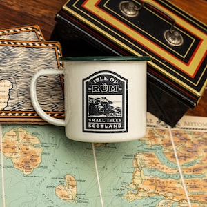 Op de afbeelding: Een witte emaille mok met een groene rand. De mok heeft een zwart-witte afbeelding van een bergketen met de tekst "Isle of Rum" en "Small Isles Scotland".