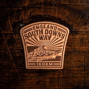 Pode incluir: Um emblema bronzeado com o texto "ENGLAND SOUTH DOWNS WAY" e uma cena de montanha. O emblema também apresenta "160KM". O design está sobre um fundo escuro e texturizado.