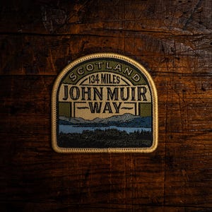 May include: A patch with the text "SCOTLAND 134 MILES JOHN MUIR WAY" in a rectangular shape with a curved top. The patch features a landscape design with a lake, mountains, and trees. The patch is on a wooden surface.