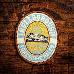 May include: Oval patch with the text "INGLEBOROUGH YORKSHIRE DALES" in blue and white. The center features a yellow sunburst design with a black and white mountain illustration and the text "723M". The patch has a brown border.
