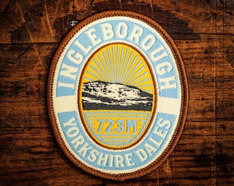 May include: Oval patch with the text "INGLEBOROUGH YORKSHIRE DALES" in blue and white. The center features a yellow sunburst design with a black and white mountain illustration and the text "723M". The patch has a brown border.