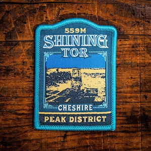 Peut inclure: Un écusson rectangulaire avec une bordure bleue. Le design présente le texte "SHINING TOR" et "PEAK DISTRICT". L'écusson comprend le texte "559M" et "CHESHIRE". Le design présente une illustration de paysage bleu et jaune.