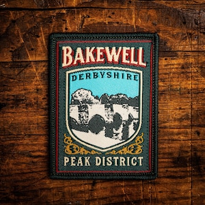 Op de afbeelding: Rechthoekige patch met de tekst "BAKEWELL DERBYSHIRE PEAK DISTRICT" in rood en wit. Het ontwerp toont een schild met een blauwe lucht en een landschapsillustratie. Donkergroene rand op houten achtergrond.