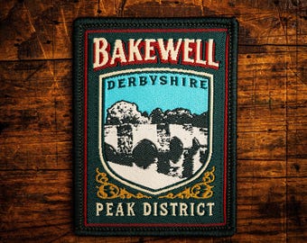 May include: A rectangular patch with the text "BAKEWELL DERBYSHIRE PEAK DISTRICT". The patch features a shield design with a blue sky and a landscape illustration. The patch has a dark green border and is set against a wooden background. The text is in red and white.