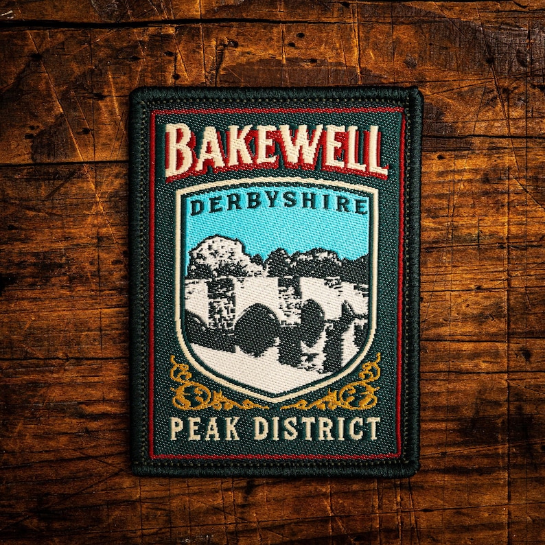 K&ouml;nnte beinhalten: Rechteckiger Aufn&auml;her mit dem Text "BAKEWELL DERBYSHIRE PEAK DISTRICT" in Rot und Wei&szlig;. Das Design zeigt einen Schild mit blauem Himmel und Landschaftsillustration. Dunkelgr&uuml;ner Rand auf Holzgrund.