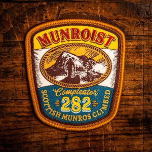 May include: A patch with the text "MUNROIST" in yellow, above an oval illustration of a mountain range. Below, it reads "Compleator 282 Scottish Munros Climbed" in blue and yellow. The patch has a brown border.