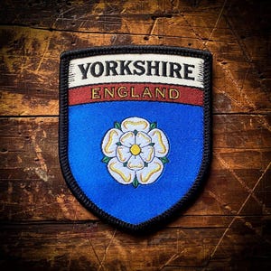 Puede incluir: Un parche bordado azul y blanco con el texto "Yorkshire England" y una rosa blanca con un centro amarillo.