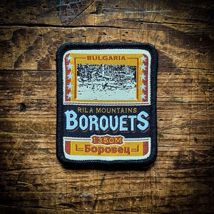 Op de afbeelding: Een zwart en geel geborduurde patch met de tekst "Borovets" in blauw, "Rila Mountains" in zwart en "Bulgaria" in wit. De patch heeft ook een zwart-wit afbeelding van een bergdorp en de tekst "1350m Borovets" in geel.