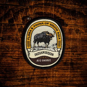 May include: An oval embroidered patch featuring a dark grey Indian bison illustration on a cream background. The border is black and gold with the text FLORA & FAUNA OF SOUTH ASIA, INDIAN BISON, and BOS GAURUS printed in black lettering.
