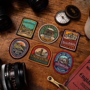 K&ouml;nnte beinhalten: Eine Sammlung gestickter Aufn&auml;her mit verschiedenen Orten der Cotswolds in England. Die Aufn&auml;her zeigen Bilder von Landschaften, Geb&auml;uden und Texten wie "Bibury" und "Broadway". Ein Kompass und eine Kamera sind ebenfalls zu sehen.