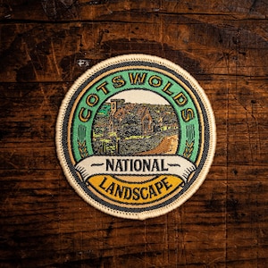 Peut inclure: Écusson rond avec l'inscription "Cotswolds National Landscape" en vert, jaune et blanc. L'écusson présente une illustration d'un bâtiment et d'arbres. Il a une bordure noire et est posé sur un fond de grain de bois foncé.