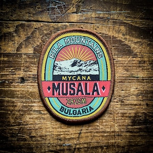 Puede incluir: Un parche circular marrón con un diseño colorido que representa una cordillera con un sol saliendo detrás. El texto "Rila Mountains" está escrito en forma circular alrededor de la parte superior del parche. El texto "Mycana Musala 2925M Bulgaria" está escrito en forma circular alrededor de la parte inferior del parche.
