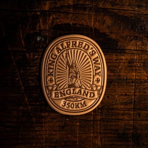 Pode incluir: Um emblema circular de cor bronze com o texto "KING ALFRED'S WAY ENGLAND" e uma representação de uma figura segurando um bastão. O emblema também apresenta a medida "350KM". O fundo é uma superfície de madeira escura e texturizada.