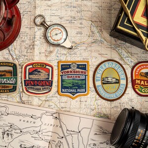 May include: A selection of Yorkshire Dales patches, a compass, and a camera are displayed on a vintage map. The patches feature text like "Whernside" and "Pen-y-Ghent" with measurements in metres. A red lantern and other travel accessories complete the scene.