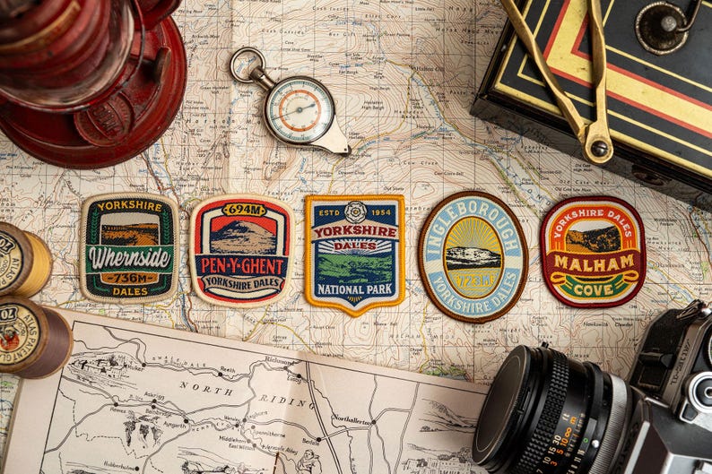 May include: A selection of Yorkshire Dales patches, a compass, and a camera are displayed on a vintage map. The patches feature text like "Whernside" and "Pen-y-Ghent" with measurements in metres. A red lantern and other travel accessories complete the scene.