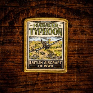 May include: A rectangular embroidered patch with a gold border. The top section features the text HAWKER TYPHOON in green. The centre shows a Hawker Typhoon aircraft flying over a landscape. The bottom section reads BRITISH AIRCRAFT OF WWII.