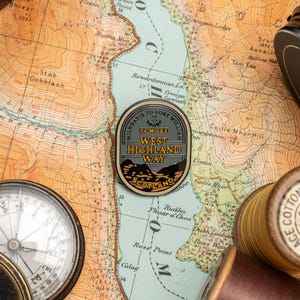 Könnte beinhalten: Ein runder Anstecker mit dem Text "Milngavie to Fort William, 96 Miles, West Highland Way, Scotland" liegt auf einer Vintage-Karte. Ein Kompass und Garnrollen sind ebenfalls zu sehen. Der Anstecker hat ein graues und schwarzes Design.