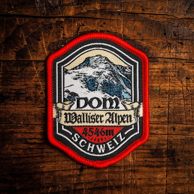 May include: A patch with a red border, featuring a mountain scene with the text "DOM Walliser Alpen 4546m SCHWEIZ". The patch has a black and white design with a mountain peak in the center.