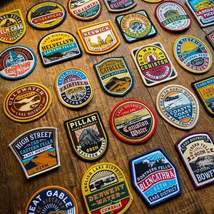 May include: A collection of colorful embroidered patches featuring various locations in the Lake District National Park in England. Each patch features a different mountain peak, lake, or town, along with the name of the location and its elevation in meters. The patches are made of a durable fabric and are perfect for adding a touch of personality to backpacks, jackets, or other items.