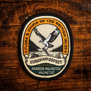 Könnte beinhalten: Ovaler Aufnäher mit schwarzem Rand, der einen Fischadler im Flug zeigt. Der Aufnäher enthält den Text "Flora & Fauna of the British Isles" und "Eurasian Osprey". Der Hintergrund ist eine Holzoberfläche, die dem Aufnäher ein rustikales Aussehen verleiht.