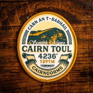 May include: An oval embroidered patch with a cream and gold design. The patch features a mountain scene in green and gold, with text that reads "Scottish Munro CAIRN TOUL 4236'" and "CAIRNGORMS". The height is 1291m.