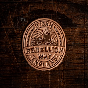 Könnte beinhalten: Ein kreisförmiges, bronzefarbenes Emblem mit dem Text "373KM" oben. Das Emblem zeigt die Worte "REBELLION WAY ENGLAND" und eine Landschaftsgrafik mit einem Sonnenstrahlendesign.