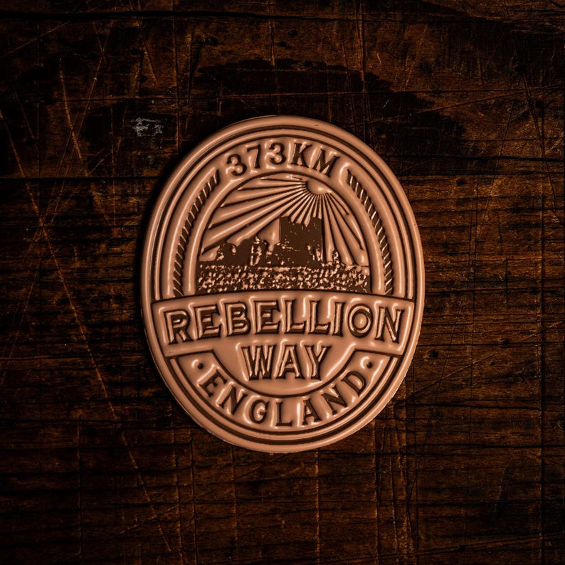 K&ouml;nnte beinhalten: Ein kreisf&ouml;rmiges, bronzefarbenes Emblem mit dem Text "373KM" oben. Das Emblem zeigt die Worte "REBELLION WAY ENGLAND" und eine Landschaftsgrafik mit einem Sonnenstrahlendesign.