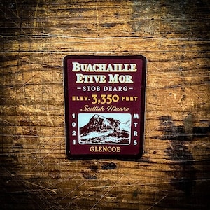 May include: A burgundy sticker with white text that reads "Buachaille Etive Mor - Stob Dearg - Elev. 3,350 Feet Scottish Munro Glencoe". The sticker features a small illustration of a mountain.