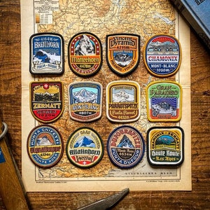 Pode incluir: Nove patches bordados representando diferentes picos de montanha e seus nomes, incluindo o Matterhorn, o Mont Blanc e o Breithorn. Os patches est&atilde;o dispostos em uma grade em um mapa vintage com um fundo de madeira.