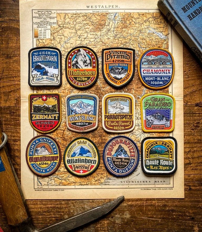 Pode incluir: Nove patches bordados representando diferentes picos de montanha e seus nomes, incluindo o Matterhorn, o Mont Blanc e o Breithorn. Os patches est&atilde;o dispostos em uma grade em um mapa vintage com um fundo de madeira.