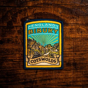 Może przedstawiać: Prostokątna naszywka z szczegółową ilustracją sceny wioski z napisem "ENGLAND BIBURY COTSWOLDS". Naszywka ma niebieską obwódkę i żółte akcenty. Tło to ciemne usłojenie drewna.