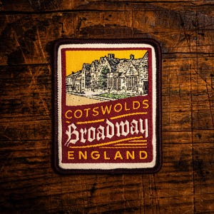 Może przedstawiać: Haftowana naszywka z ilustracją budynków i napisem "Cotswolds Broadway England". Naszywka ma prostokątny kształt z bordową obwódką, żółtym tłem i kremową wewnętrzną obwódką. Tekst jest w stylizowanej czcionce.