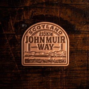 Pode incluir: Uma placa de metal cor de bronze com o texto "SCOTLAND 215KM JOHN MUIR WAY" em forma semicircular. A placa apresenta uma ilustração de paisagem na parte inferior. A placa está sobre um fundo de madeira escura.