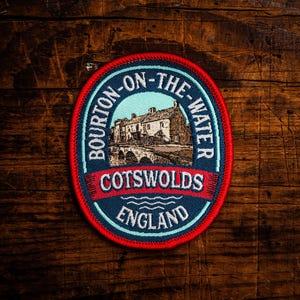 Może przedstawiać: Okrągła naszywka z czerwoną obwódką i niebiesko-białym wzorem. Naszywka zawiera napis "Bourton-on-the-Water Cotswolds England" wokół ilustracji budynku i mostu. Naszywka jest umieszczona na tle ciemnego usłojenia drewna.